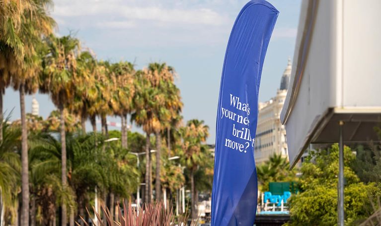 This image was captured during the Cannes Lions International Festival of Creativity. It features a large, blue feather flag prominently displaying the text What's your next brilliant move?