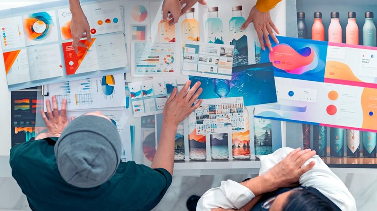 A team of designers gathered around a table, reviewing colorful mockups and data visualizations. The bird's-eye view captures their collaborative effort in a bright and modern workspace.