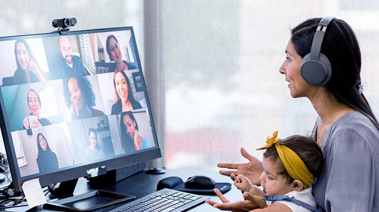 Mother, holding her baby, sits at desk while video conferencing with her co-workers.
