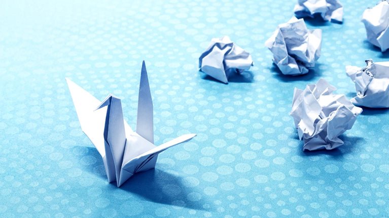 A lone origami crane sits on a blue background with white polka dots. Behind the crane is a cluster of crumpled balls of white paper.