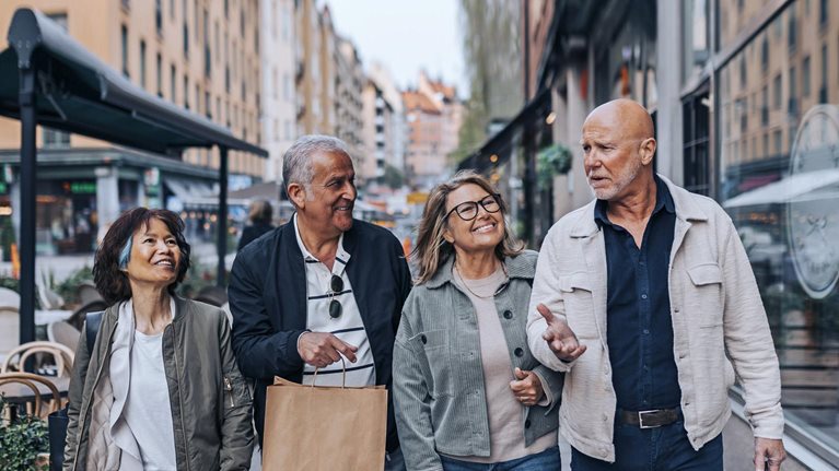 A cheerful group of senior friends strolling down a European sidewalk, engaged in conversation and carrying shopping bags.