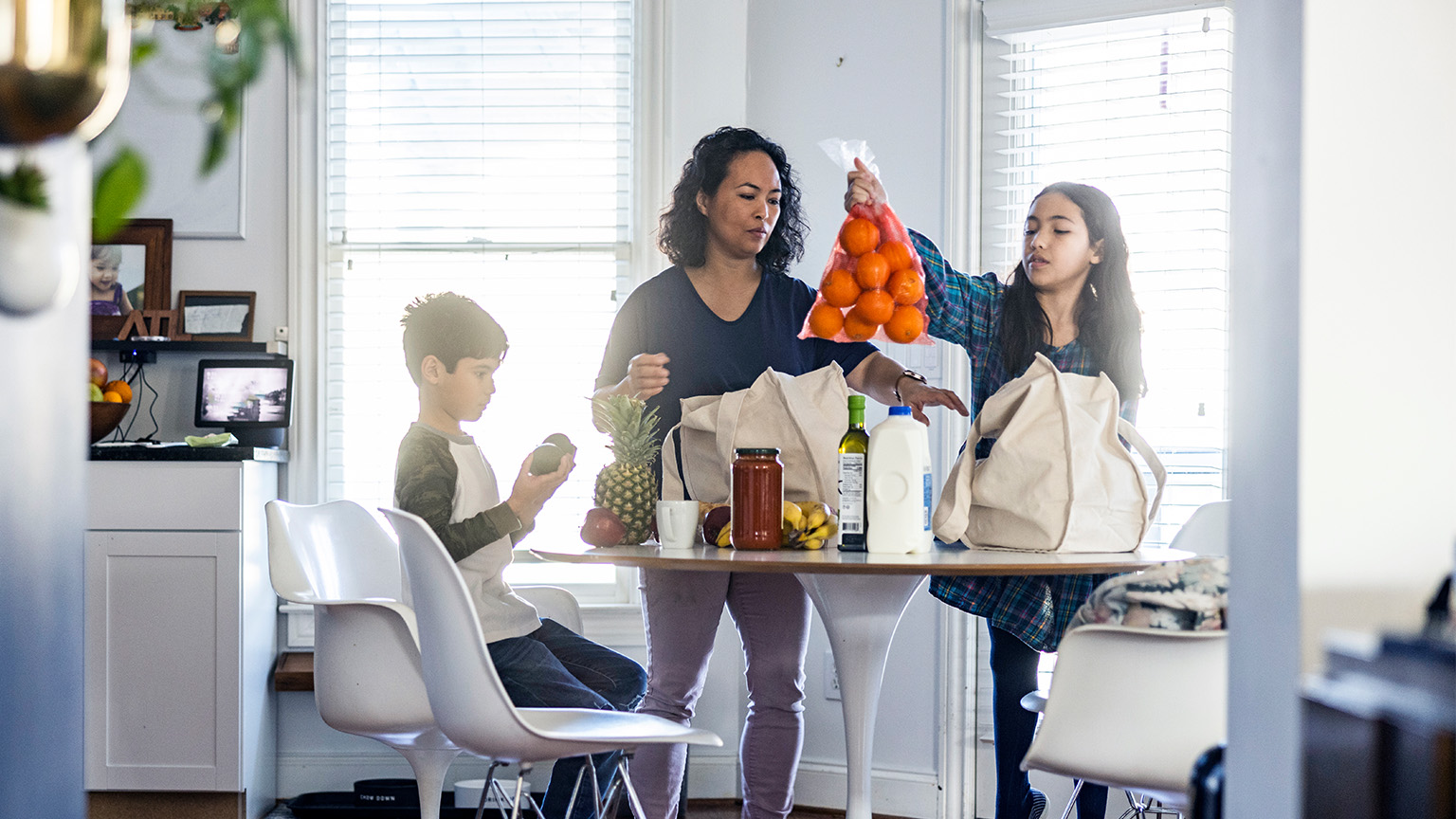 A mother is unpacking groceries at a contemporary pedestal table with her two children. Her daughter picks up a bag of oranges while her son examines some plums.