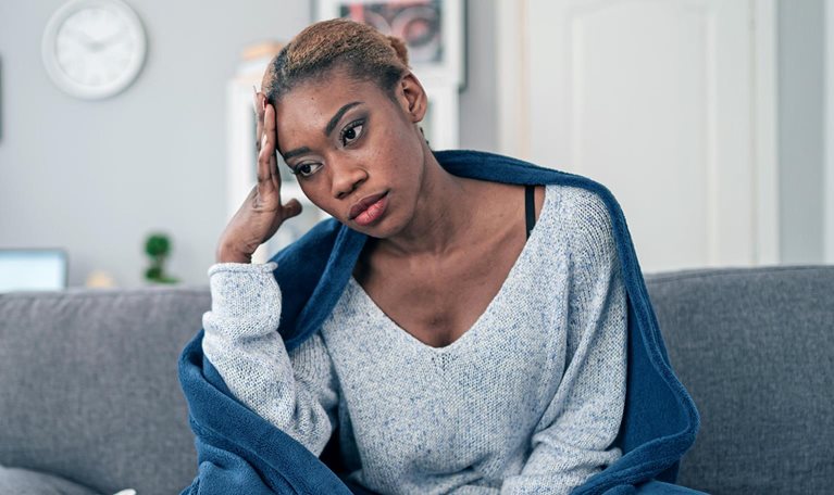 A young woman sitting on her couch wrapped in a blanket with used tissues laying next to her.