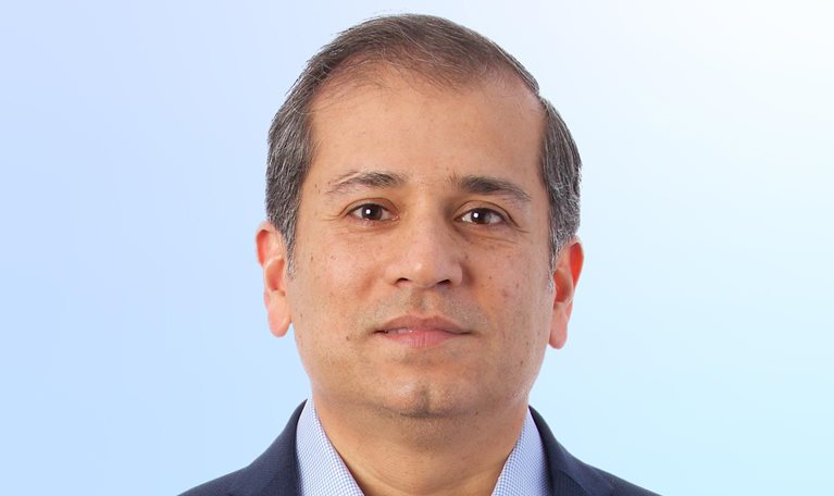 A head-and-shoulders portrait of a Shubham Singhal facing forward with a neutral expression. He is wearing a dark blazer over a light blue patterned dress shirt, set against a soft gradient background that transitions from light gray to pale blue.