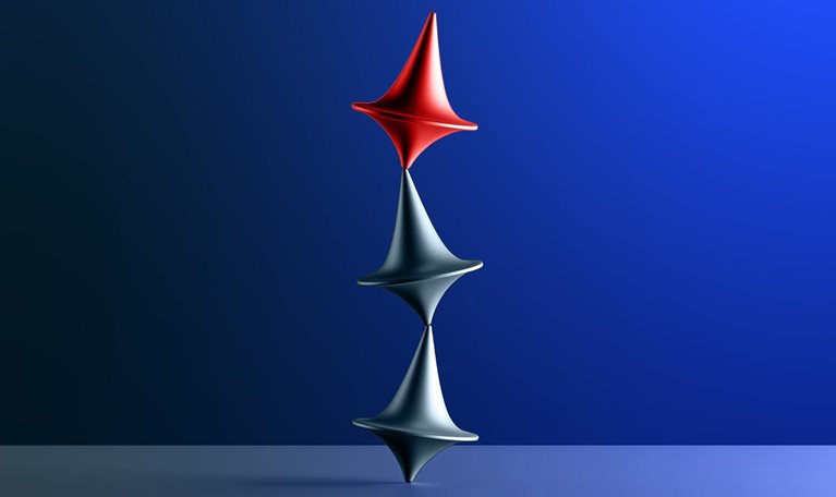 Three tops, two silver and one red, are arranged in a vertical stack on a blue surface. The tops are uneasily balanced, with the red top positioned above the two silver ones.